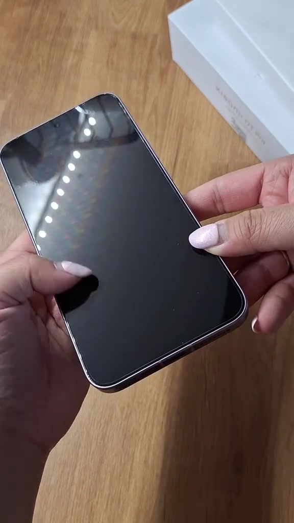 Dua tangan memegang Xiaomi 17T Pro dari depan dengan skrin hitam berkaca, kotak putih kelihatan di latar belakang atas meja kayu