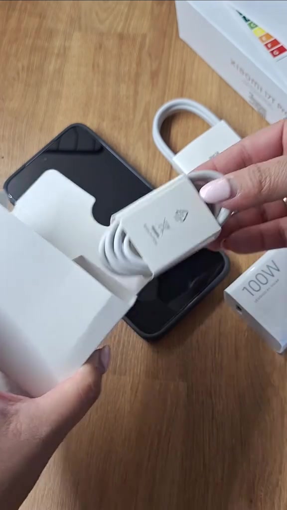 Tangan memegang kabel putih berbungkus dengan kotak adapter bertulis 100W di sebelah kanan, telefon terbaring atas meja kayu
