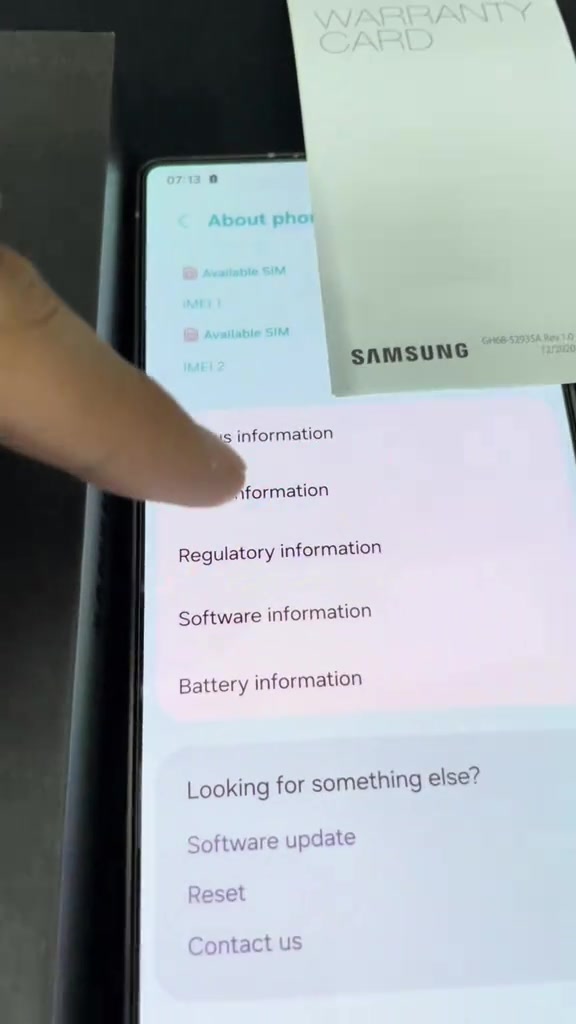 Kad waranti Samsung diletakkan sebelah skrin About phone yang menunjukkan maklumat IMEI dan slot SIM