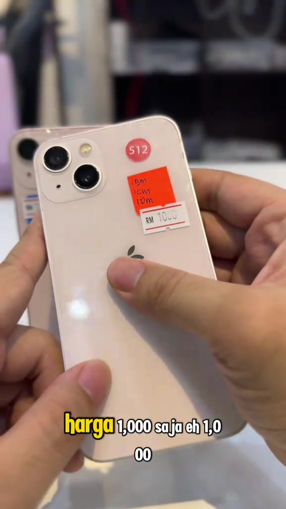 Bahagian belakang iPhone 13 warna pink dipegang tangan, ada pelekat harga tulisan tangan RM 1000 dan pelekat bulat 512