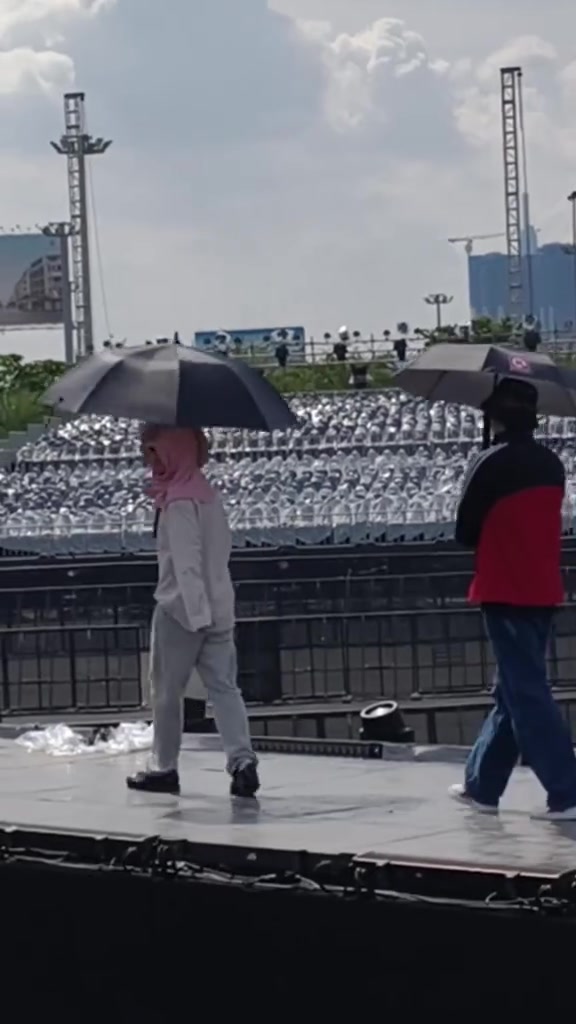 Hai người che dù đi trên sàn sân khấu ngoài trời trước khán đài trống giữa trưa nắng