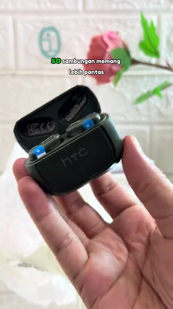 Tangan memegang case pengecas HTC hitam terbuka dengan dua earbud HTC NE51 di dalamnya