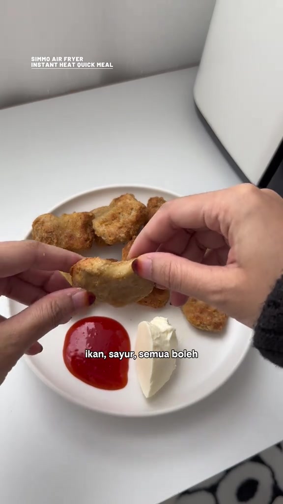 Tangan memegang nugget goreng air fryer di atas pinggan putih bersama sos cili dan mayonis