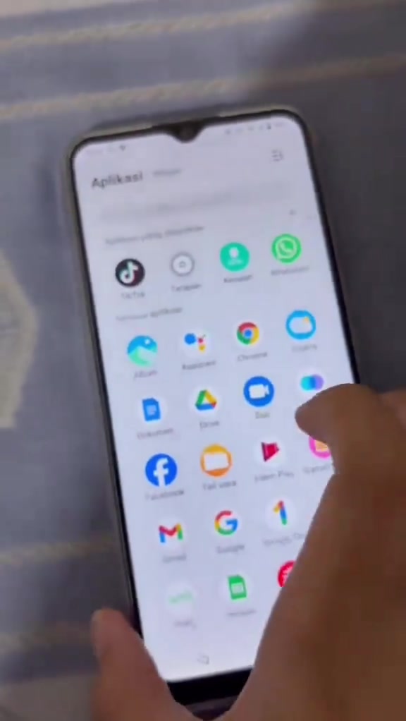 Tangan memegang telefon pintar dengan skrin senarai aplikasi, keadaan luar nampak biasa seperti phone asli