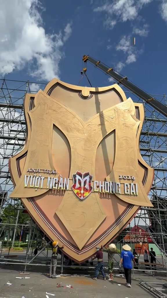 Cổng chào hình khiên in dòng chữ Anh Trai Vượt Ngàn Chông Gai 2026 đang được cần cẩu dựng lên giữa giàn giáo, vài người thợ đội nón làm việc phía dưới, nhìn từ phía sau