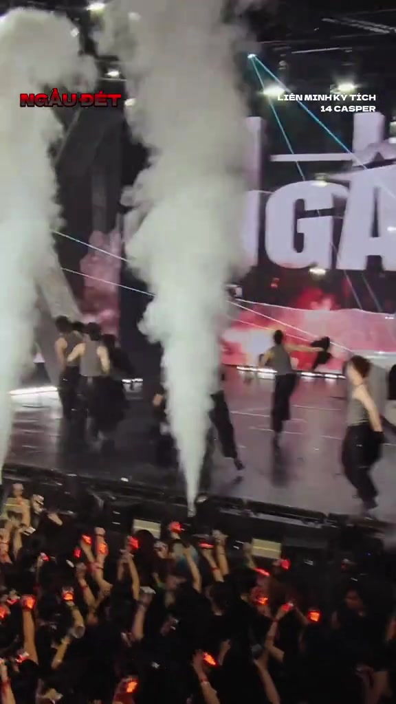 Sân khấu đêm diễn phun khói trắng với vũ công tập diễn, phía dưới là hàng khán giả giơ lightstick nhìn từ phía sau