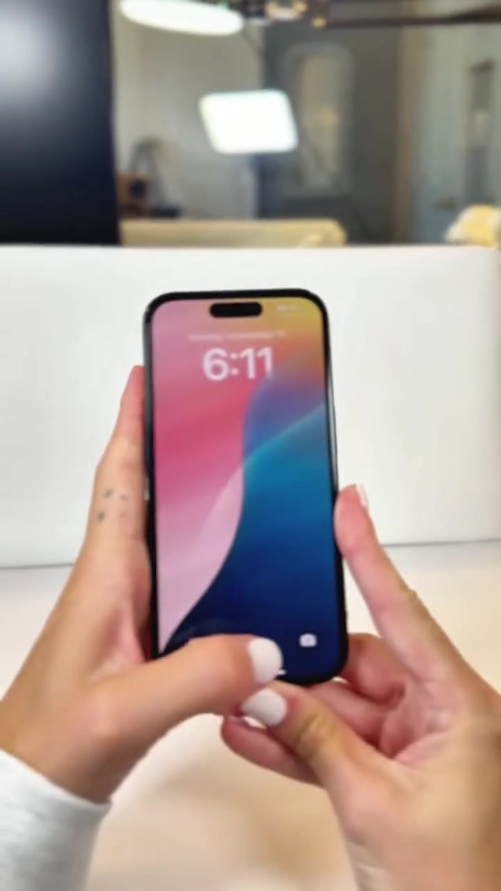 Sepasang tangan memegang iPhone hitam dengan skrin kunci menyala di atas meja putih, unit refurbished selepas pemeriksaan
