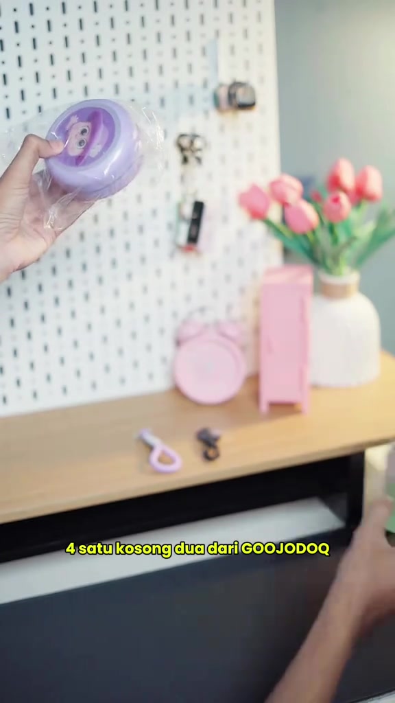 Tangan memegang speaker bulat Goojodoq warna ungu masih berbalut plastik di depan meja dengan aksesori merah jambu