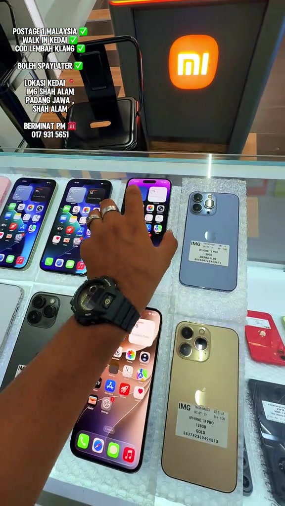 Tangan menunjuk iPhone 13 Pro warna Sierra Blue berlabel 256GB dalam cermin pameran kedai telefon terpakai