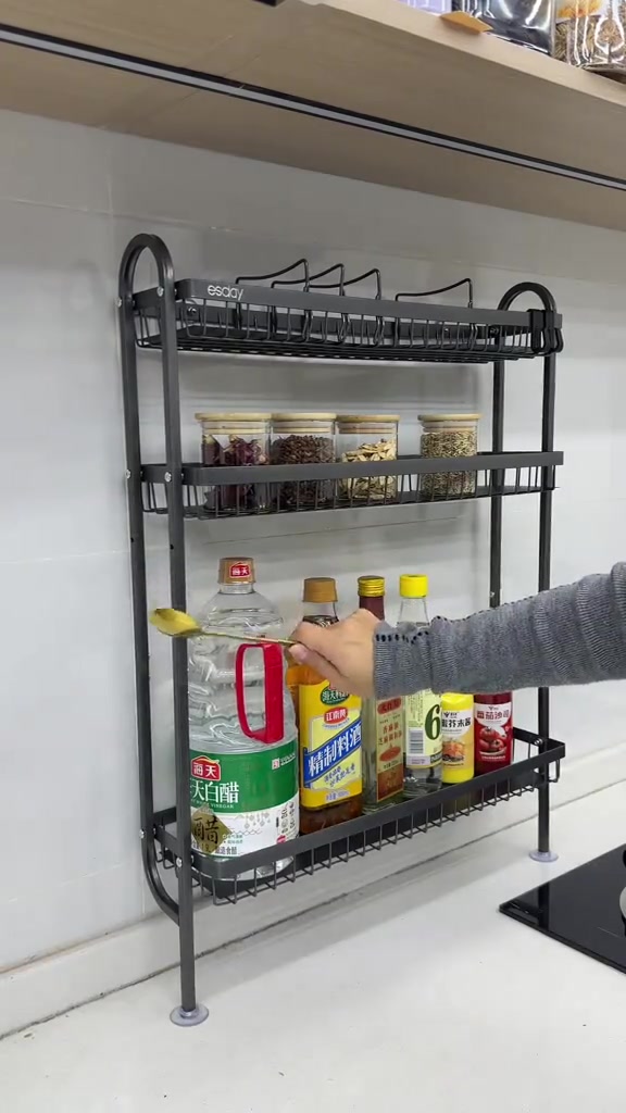 Rak rempah besi tiga tingkat lekat dinding berisi balang rempah dan botol sos, tangan memegang sudu