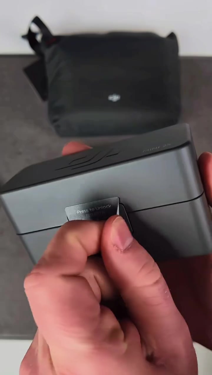 Bekas pengecas DJI Mic Mini 2S warna kelabu gelap dipegang di tangan, dengan pouch hitam di latar belakang