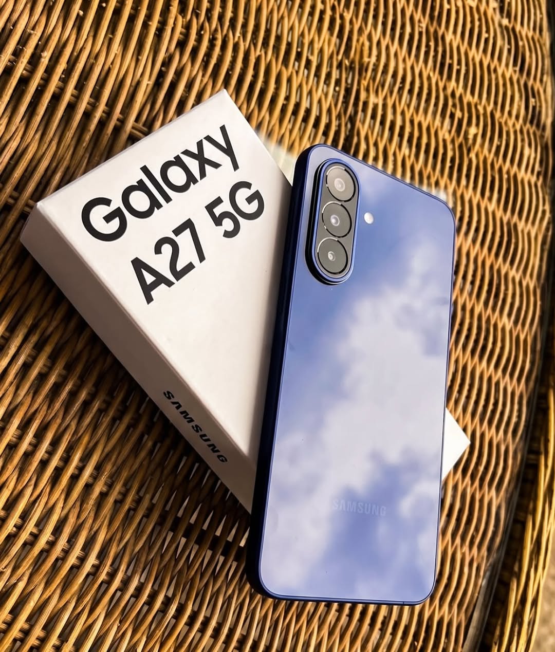 Mặt lưng màu xanh của Samsung Galaxy A27 5G với cụm ba camera đặt cạnh hộp giấy in chữ Galaxy A27 5G trên mặt mây đan