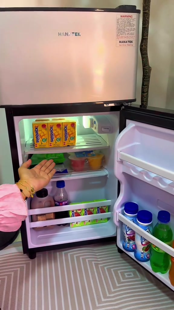 Bahagian dalam peti sejuk mini dua pintu terbuka dengan rak pintu berisi botol minuman dan tin, tangan menunjuk ke dalam ruang fridge