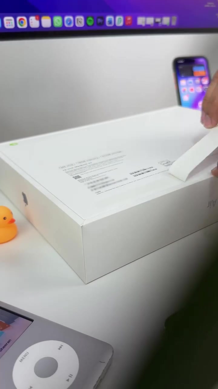 Hộp MacBook Air M5 còn niêm phong đặt trên bàn trắng, phía sau là một chiếc iPhone