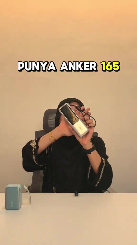 Powerbank Anker perak dengan kabel tergantung dipegang diangkat, di meja sebelah kiri ada powerbank Nano biru kecil