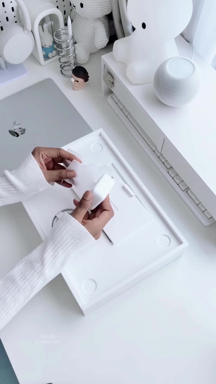 Hai tay cầm củ sạc màu trắng phía trên khay hộp MacBook Air M5, nắp máy màu bạc đặt bên cạnh