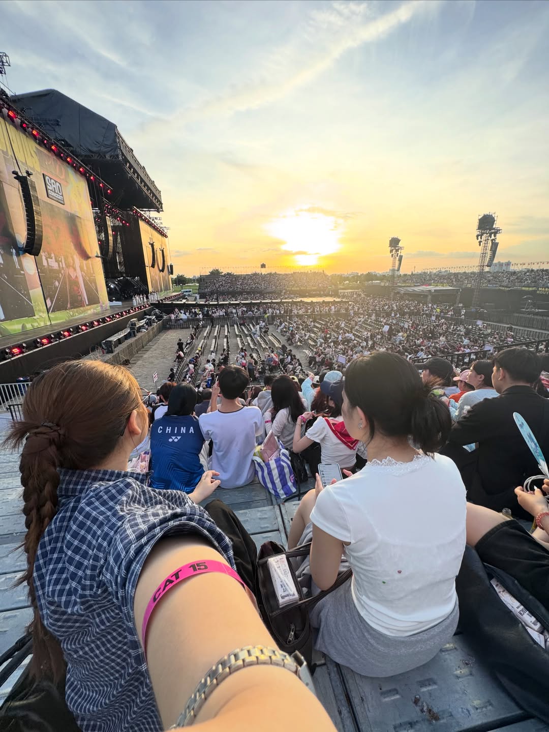 Khán đài ngồi hạng CAT nhìn về sân khấu chính SAO Concert lúc hoàng hôn, tiền cảnh là cánh tay đeo vòng tay hạng vé màu hồng, khán giả ngồi quay lưng về ống kính
