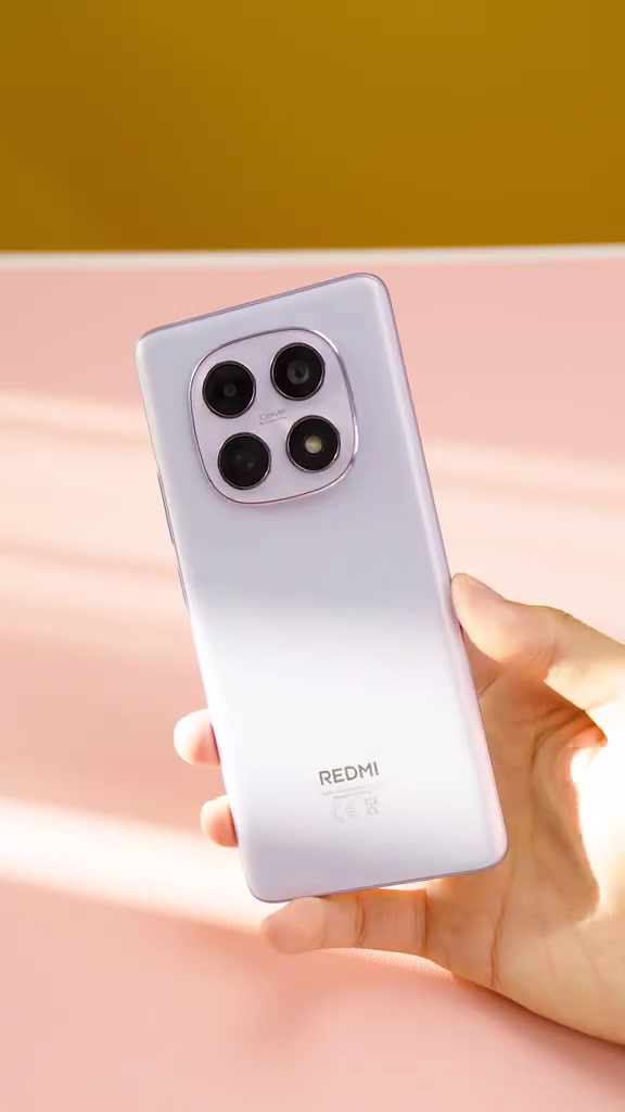 Mặt lưng màu tím nhạt của Redmi Note 15 với cụm bốn camera và logo REDMI, cầm trên tay nền hồng