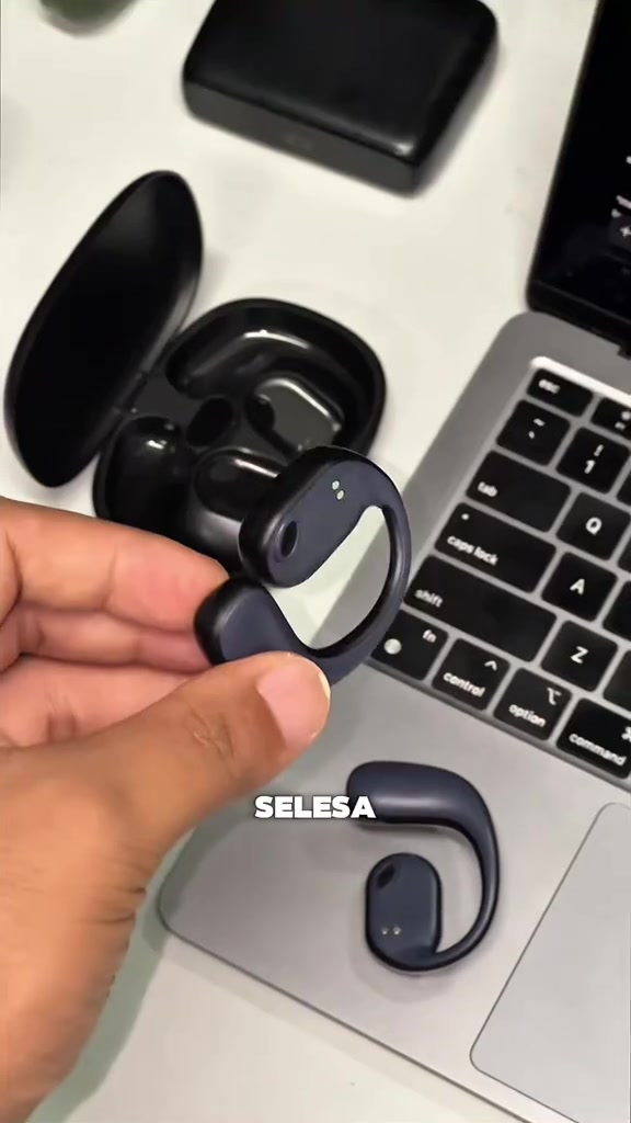 Earbud klip open-ear warna hitam dipegang di jari, dengan kes pengecas terbuka dan satu unit lagi di atas riba komputer riba