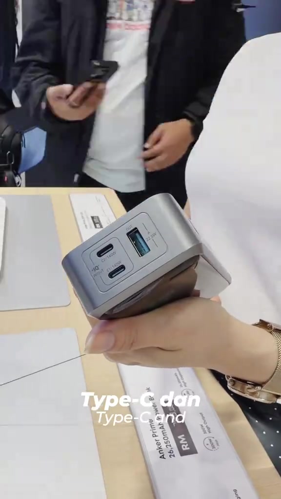 Tangan memegang power bank Anker Prime berwarna kelabu dengan port USB-C dan USB-A di meja pameran kedai
