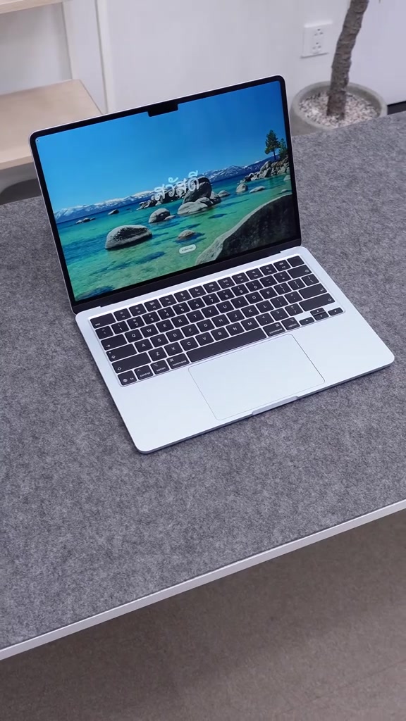 MacBook Air M5 mở sẵn trên mặt bàn phủ nỉ xám, màn hình hiện hình nền hồ nước