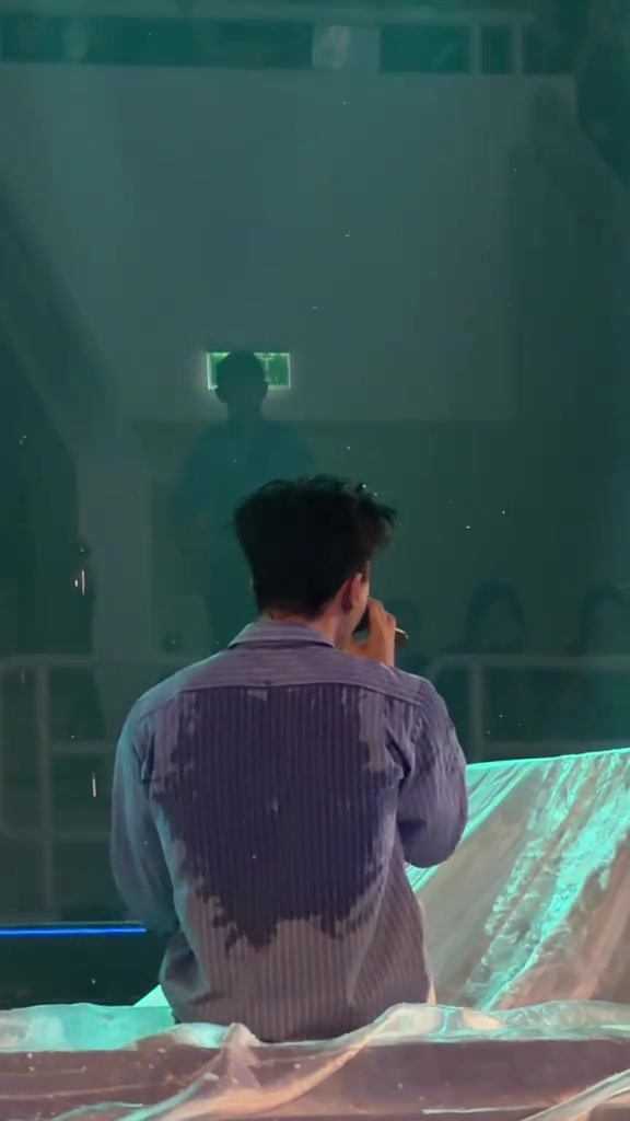Nghệ sĩ nhìn từ phía sau lưng đứng trên sân khấu concert Mắt Nhắm Mắt Mở với áo sơ mi thấm mồ hôi, hướng về màn hình và khán phòng tối
