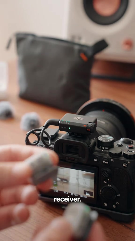 Receiver DJI Mic Mini 2S dipasang pada hot shoe kamera dengan transmitter dipegang di hadapan, pouch di latar belakang