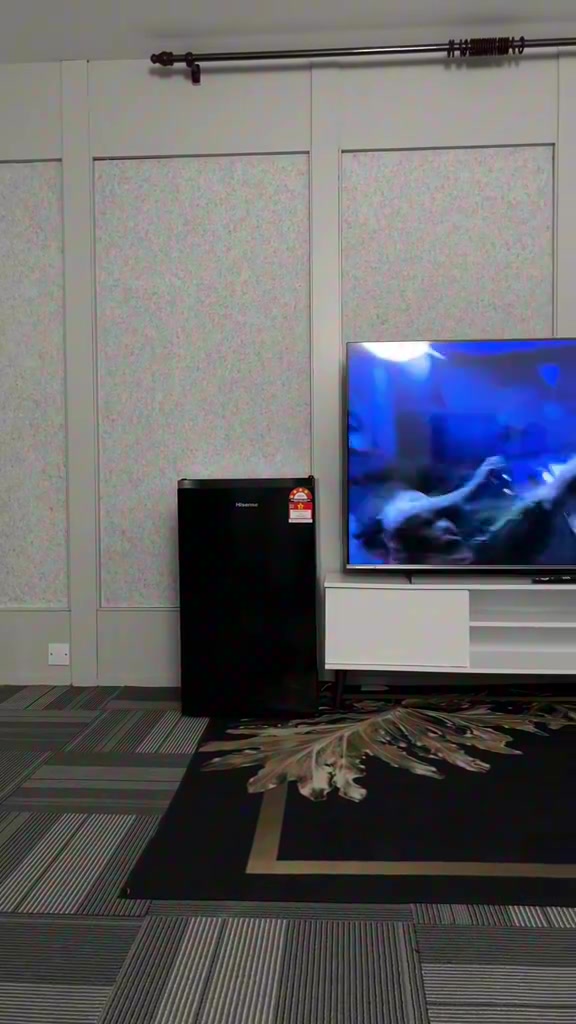 Peti sejuk mini hitam berdiri di tepi dinding bilik sebelah rak TV, menunjukkan saiznya yang tidak makan ruang
