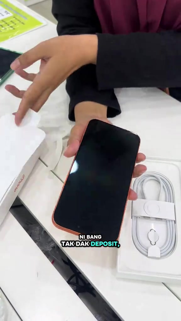 Tangan mengeluarkan iPhone berwarna oren daripada kotak bersama kabel caj putih di atas kaunter