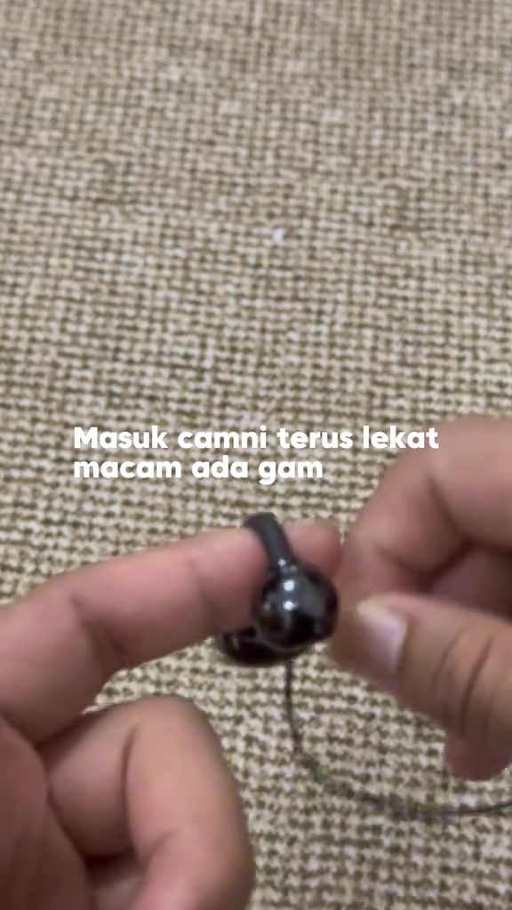 Jari memegang earbud klip kecil warna hitam jenis berwayar di atas latar kain kasar