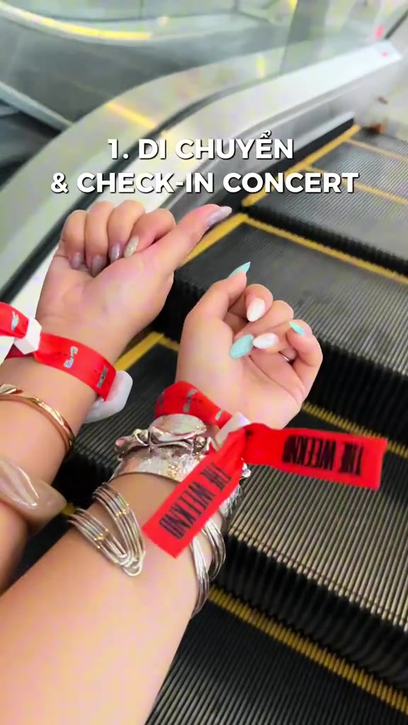 Hai cổ tay đeo vòng tay vải đỏ in chữ The Weeknd đặt trên thang cuốn lúc check-in concert ở Singapore