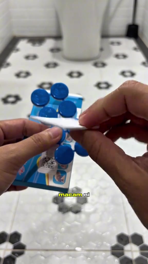 Tangan memegang kepingan umpan lipas bulat berwarna biru sambil membuka jalur double tape di belakangnya