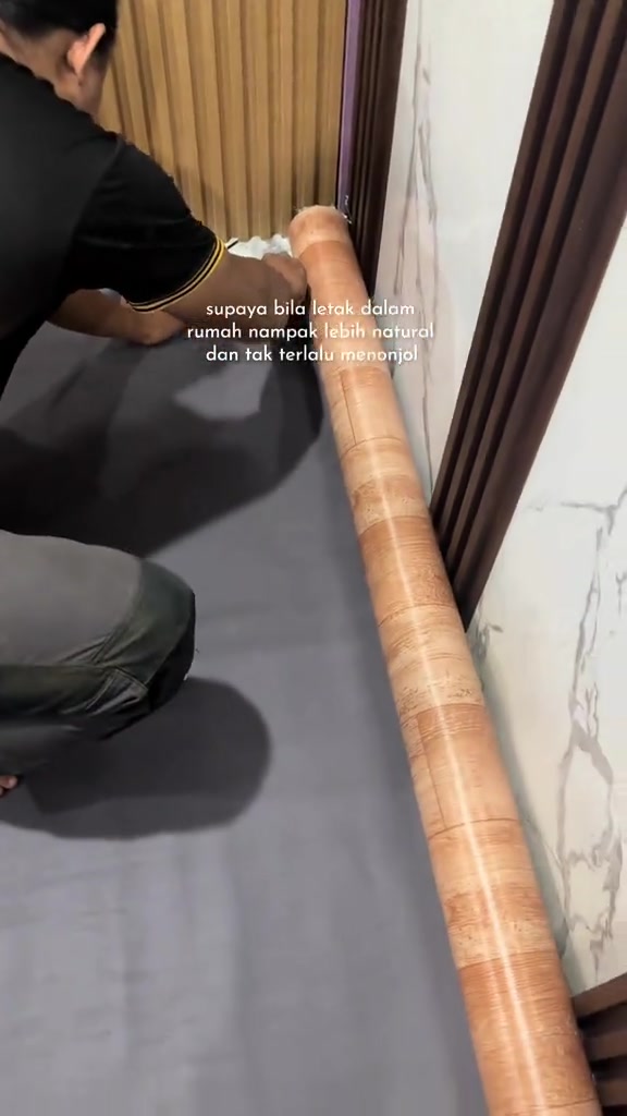 Seorang lelaki mencangkung menyelipkan tepi gulungan tikar getah rapat ke kaki dinding, tampak dari belakang