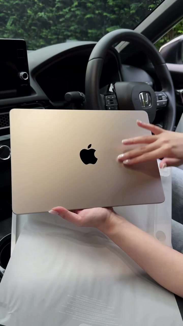 MacBook Air berwarna emas dipegang dengan satu tangan di dalam kereta, logo Apple kelihatan pada penutup