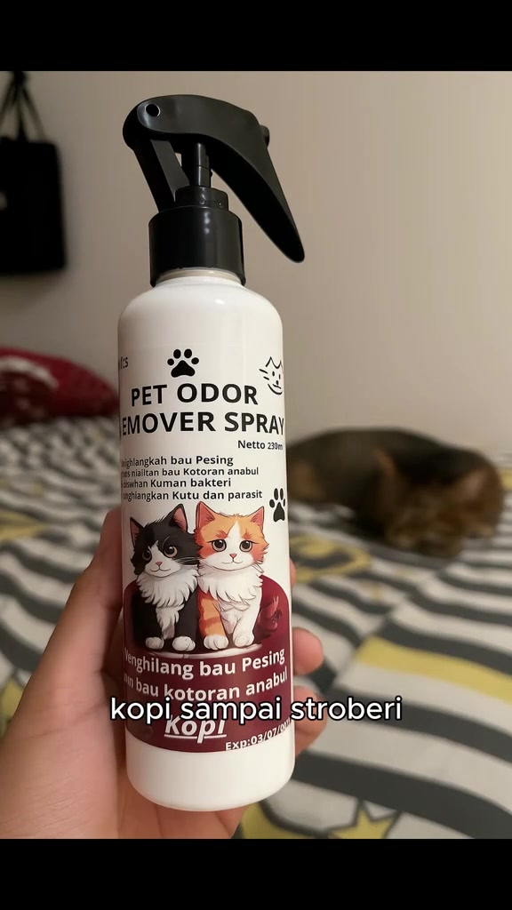 Tangan memegang botol semburan penghilang bau haiwan peliharaan 230 ml dengan seekor kucing tidur di atas katil di latar belakang
