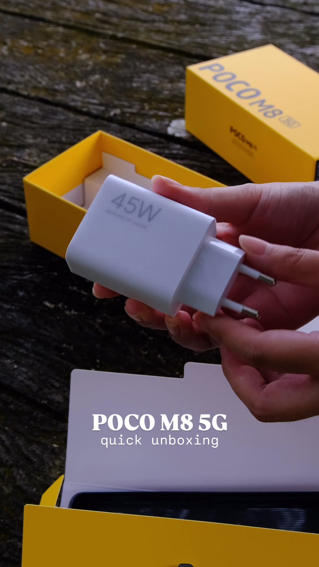 Tangan memegang pengecas putih bertanda 45W yang dikeluarkan daripada kotak POCO M8 5G.