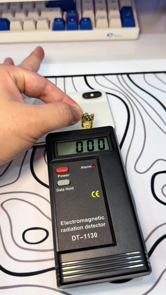 Máy đo màu đen in chữ Electromagnetic radiation detector DT-1130 hiện số 000, đặt cạnh chiếc iPhone trắng có miếng chip mạ vàng hình mèo