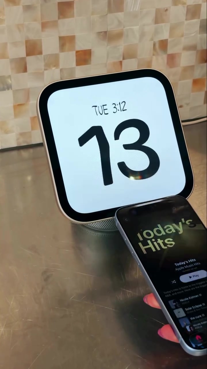 Apple HomeView bản để bàn trên mặt bàn kim loại, màn hình hiện mặt đồng hồ số lớn, phía trước có một chiếc iPhone đang phát nhạc