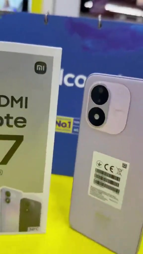 Kotak Redmi Note 17 5G berdiri sebelah unit phone warna ungu di atas meja kuning dengan latar belakang biru telco