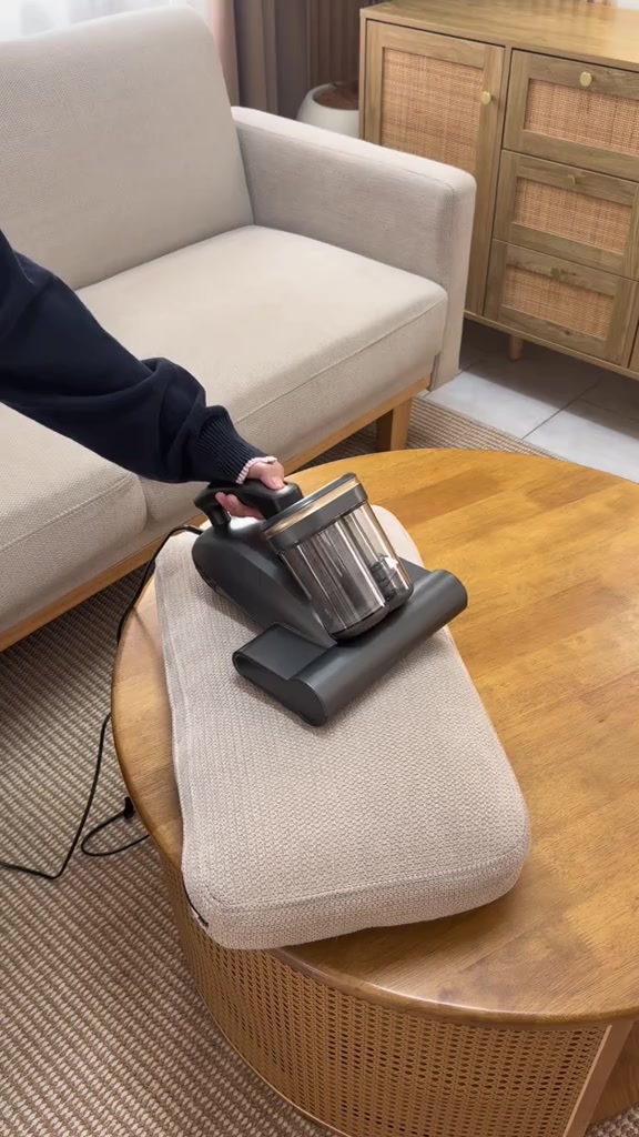 Dust mite vacuum Simplus warna kelabu gelap atas kusyen sofa di meja bulat kayu, tangan memegang pemegang