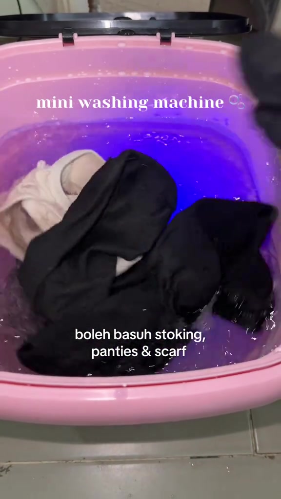 Mesin basuh mini lipat merah jambu berisi baju hitam dan coklat dengan lampu biru menyala dalam air