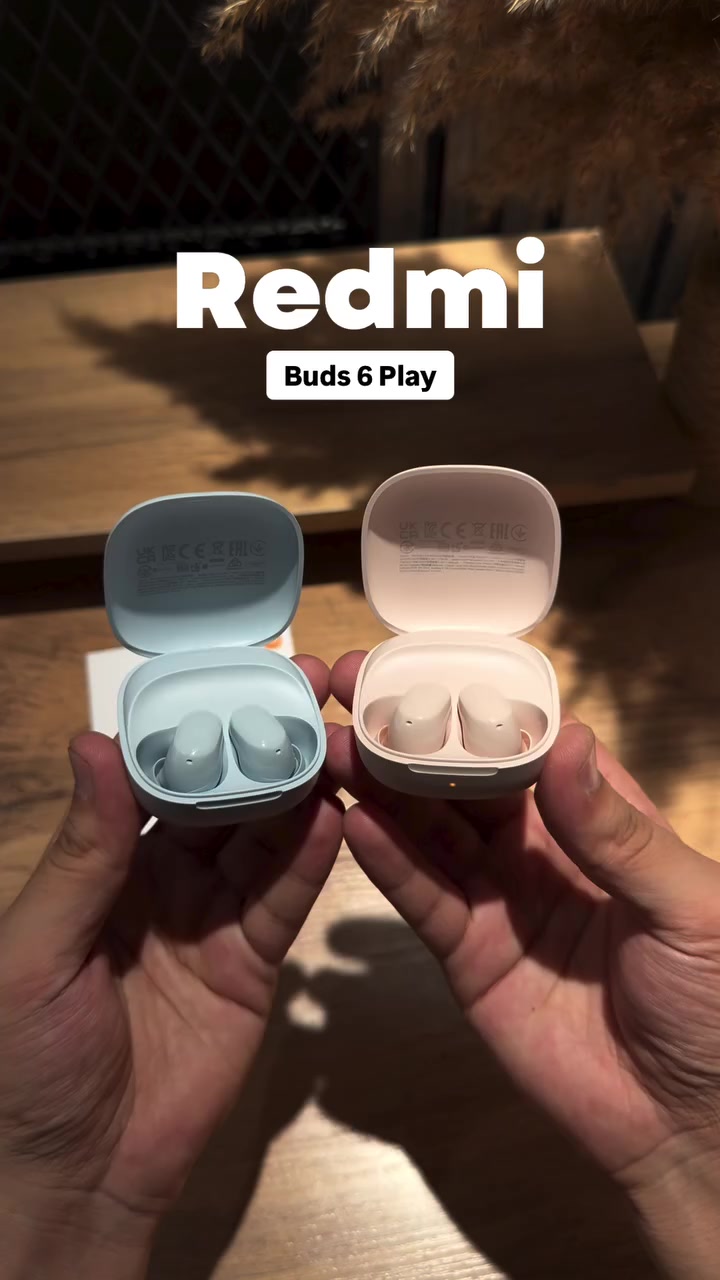 Dua tangan memegang sepasang kasus Redmi Buds 6 Play yang terbuka, warna biru muda dan merah jambu pucat.