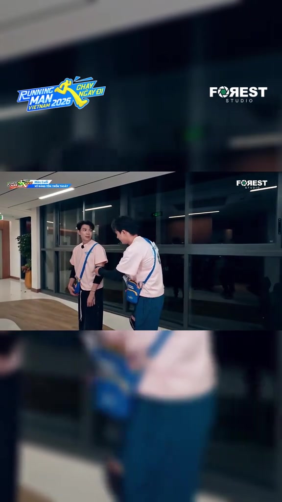 Hành lang kính trường quay buổi tối với hai thành viên áo hồng phấn đeo túi chéo xanh, một người quay lưng