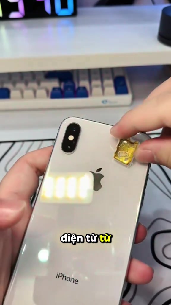 Bàn tay đang dán miếng chip mạ vàng lên mặt lưng màu trắng của một chiếc iPhone, phía sau là bàn phím cơ