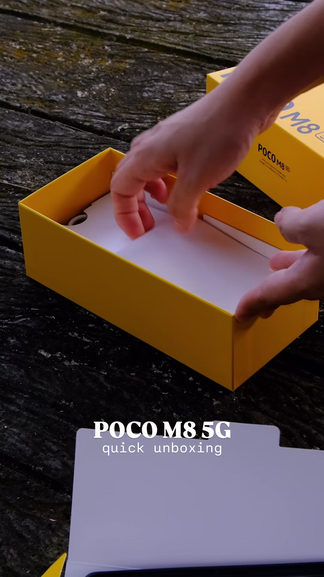 Tangan membuka kotak kuning POCO M8 5G semasa unboxing, dengan telefon masih berlapis pelindung putih di dalam kotak.