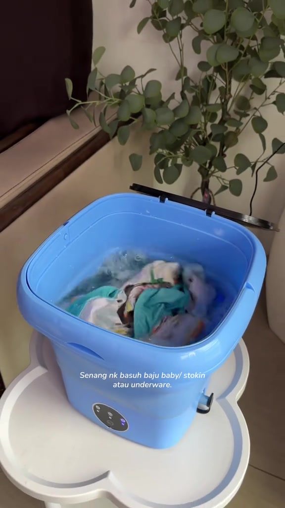 Mesin basuh mini tab biru berisi air dan baju baby diletak atas stool putih dalam ruang tamu