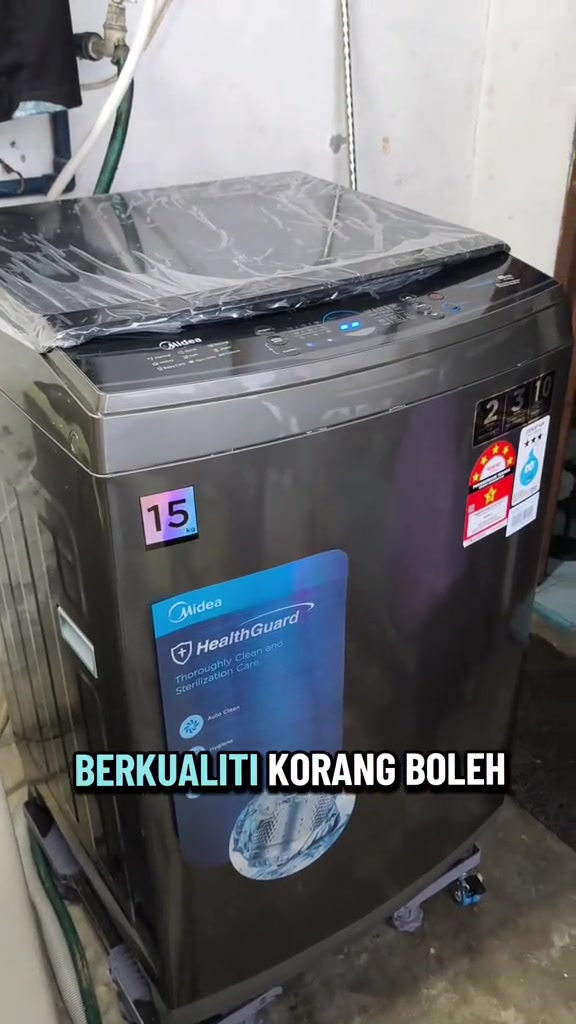 Mesin basuh top load Midea 15kg berwarna gelap masih berbalut plastik di ruang dobi rumah