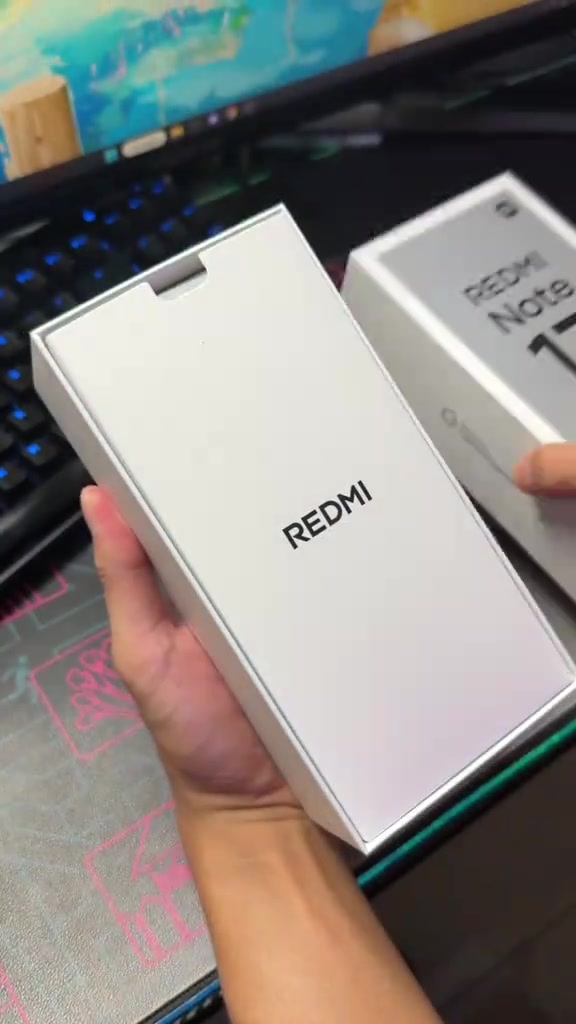 Tangan memegang kotak putih REDMI Note 17 Pro Max di atas meja dengan papan kekunci di latar
