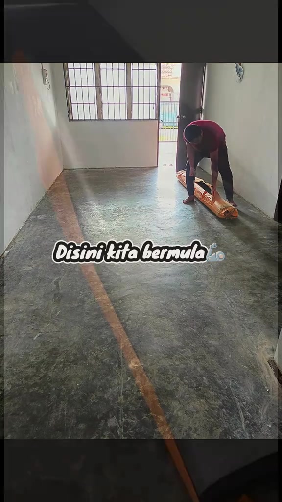 Seorang lelaki membongkok membuka gulungan tikar getah di atas lantai simen rumah sewa, tampak dari belakang
