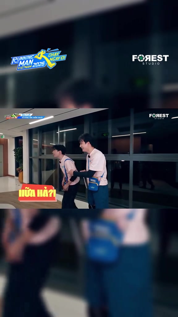 Hai thành viên áo hồng phấn đứng nghiêng đối diện nhau trong hành lang kính trường quay buổi tối